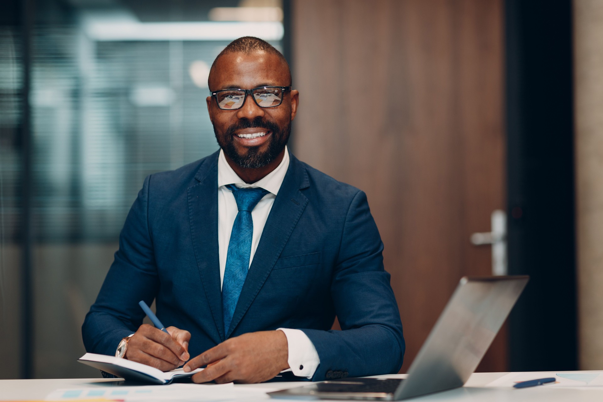 Portrait smiling african american businessman in blue suit sit at table for meeting in office with notebook with pen and laptop.